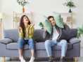 Two people sitting on gray couch covering their ears from loud noise