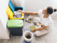Young woman sits on the floor near her trash bins to separate plastics in an effort to lead a more eco-friendly lifestyle.