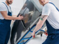 Cropped view of two movers using hand truck while transporting refrigerator in apartment.