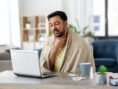 Sick man wears a blanket, drinks a hot tea, and holds his throat while researching symptoms online.