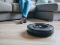 Robot vacuum cleaner zooms by a person's feet to clean hardwood floors.