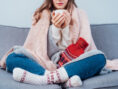 Woman keeps warm in her apartment with the help of a cozy blanket, thick socks, and a piping hot cup of tea.